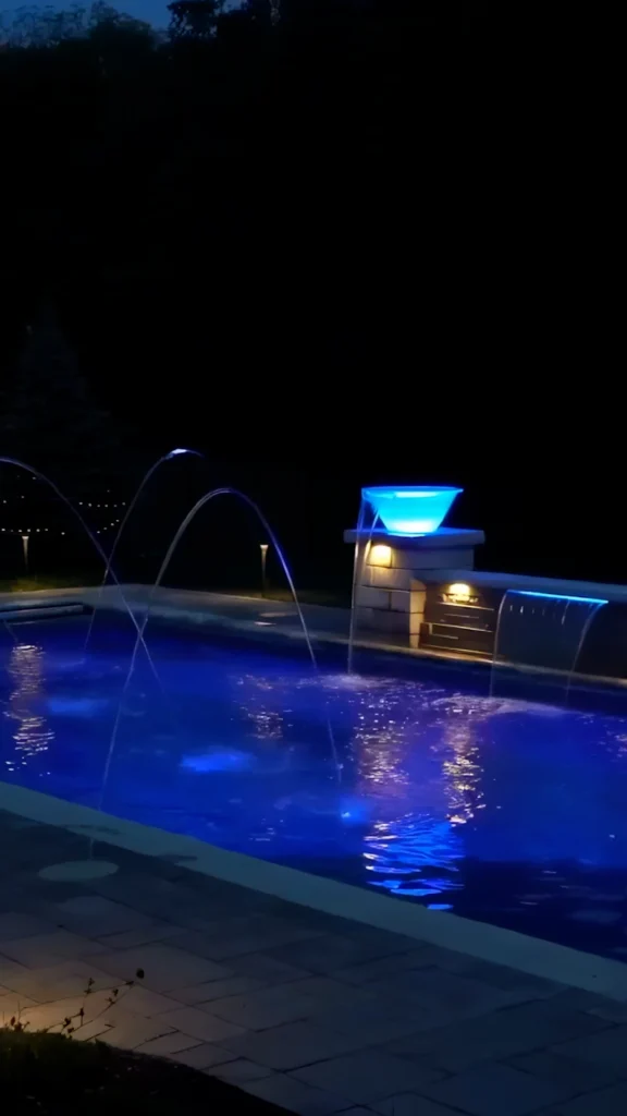 Deck-jet arcs and LED-blue scupper bowl on stacked-stone pier spilling over sheer descent into pool at night — custom pool design and installation in Alpharetta, GA by Primetime Pools.