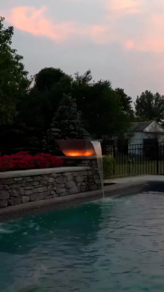 Twilight closeup of brushed stainless sheer descent water feature above natural Tennessee fieldstone wall with blooming azaleas, pastel sky — custom pool design and installation in Cumming, GA by Primetime Pools.