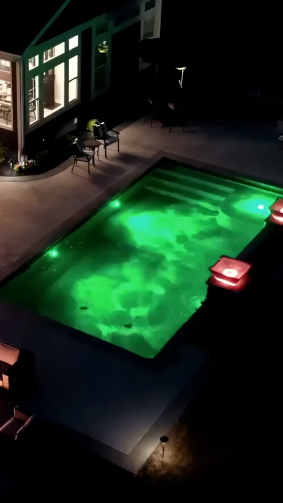 nighttime illuminated pool with LED green interior, chairs, patio decor at window-lit home — custom pool design and installation in Dacula, GA by Primetime Pools.