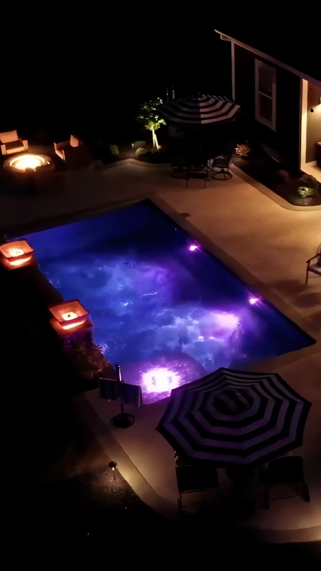 Night aerial of rectangular pool with color-changing purple LED glow, two cast-stone fire bowls on a raised stacked-stone wall, and striped umbrellas, Marietta, GA.