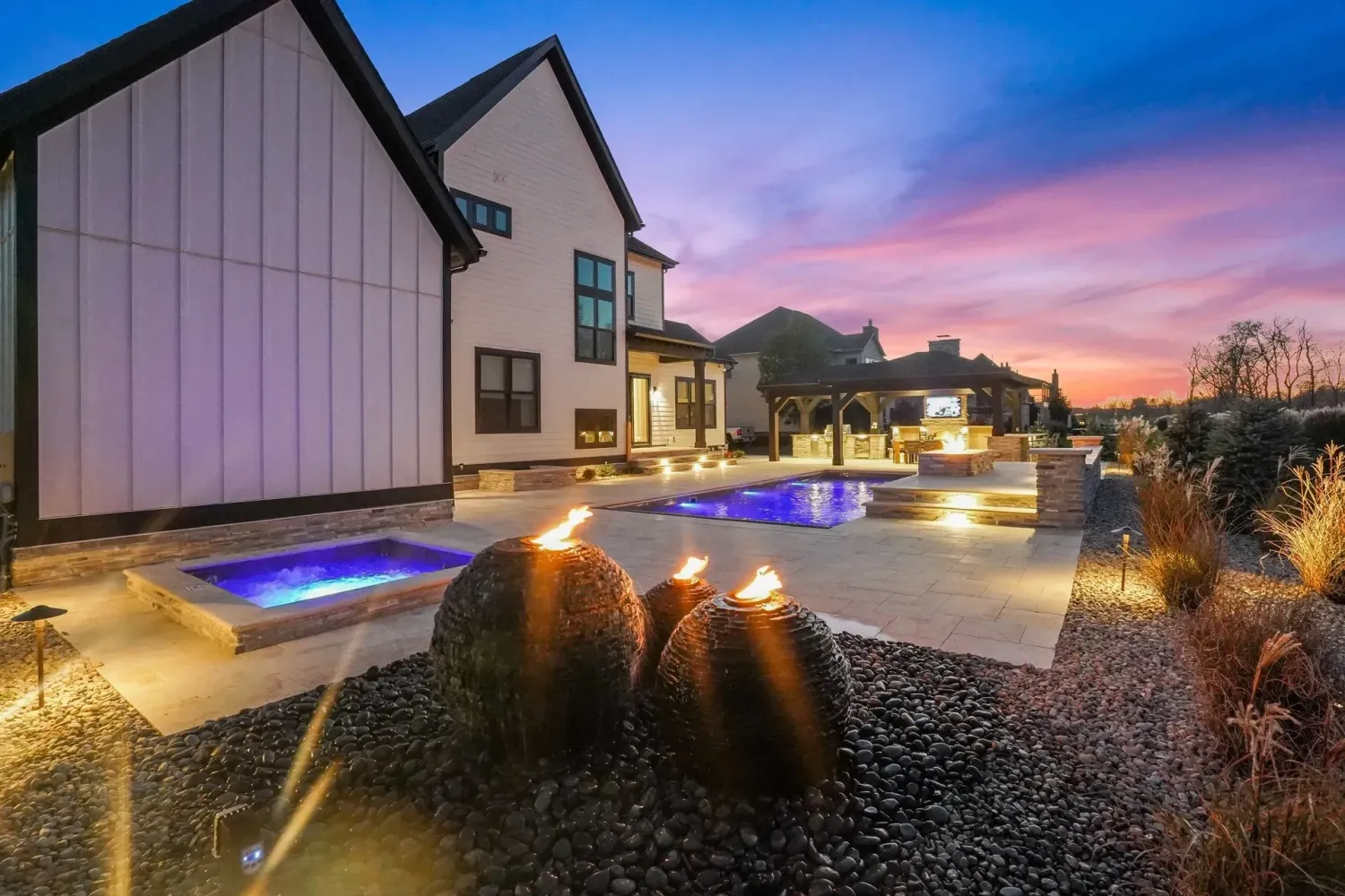 Modern farmhouse sunset hero with LED-lit pool, spa spillover, twin bronze gas fire bowls on river rock landscape and cream travertine deck, Marietta GA