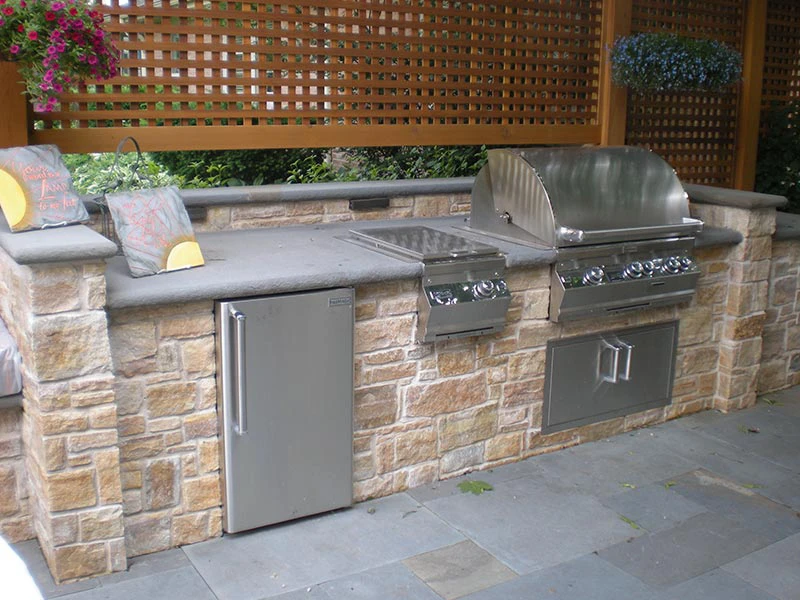 U-shape outdoor kitchen with built-in gas grill hood raised, stainless under-counter refrigerator, stacked stone base, and lattice privacy screens on a flagstone patio