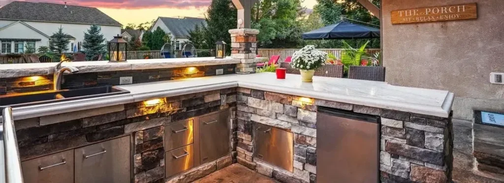 Sunset U-shape outdoor kitchen with stacked stone base, stainless drawers, carriage lanterns, 'The Porch Sit Relax Enjoy' sign — outdoor kitchen in Forsyth County, GA by Primetime Pools.