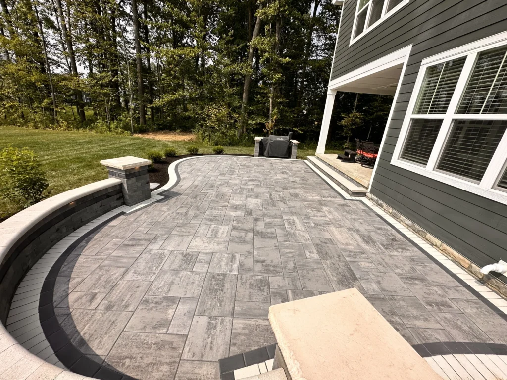 Curved gray plank paver patio with seat wall and column pier against dark gray two-story house — paver patio in Milton, GA by Primetime Pools.