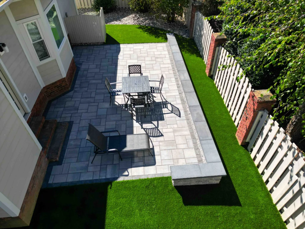 Narrow side-yard paver patio between a home and a white privacy fence, with bistro dining set and artificial turf borders — paver hardscape in Cumming, GA by Primetime Pools.