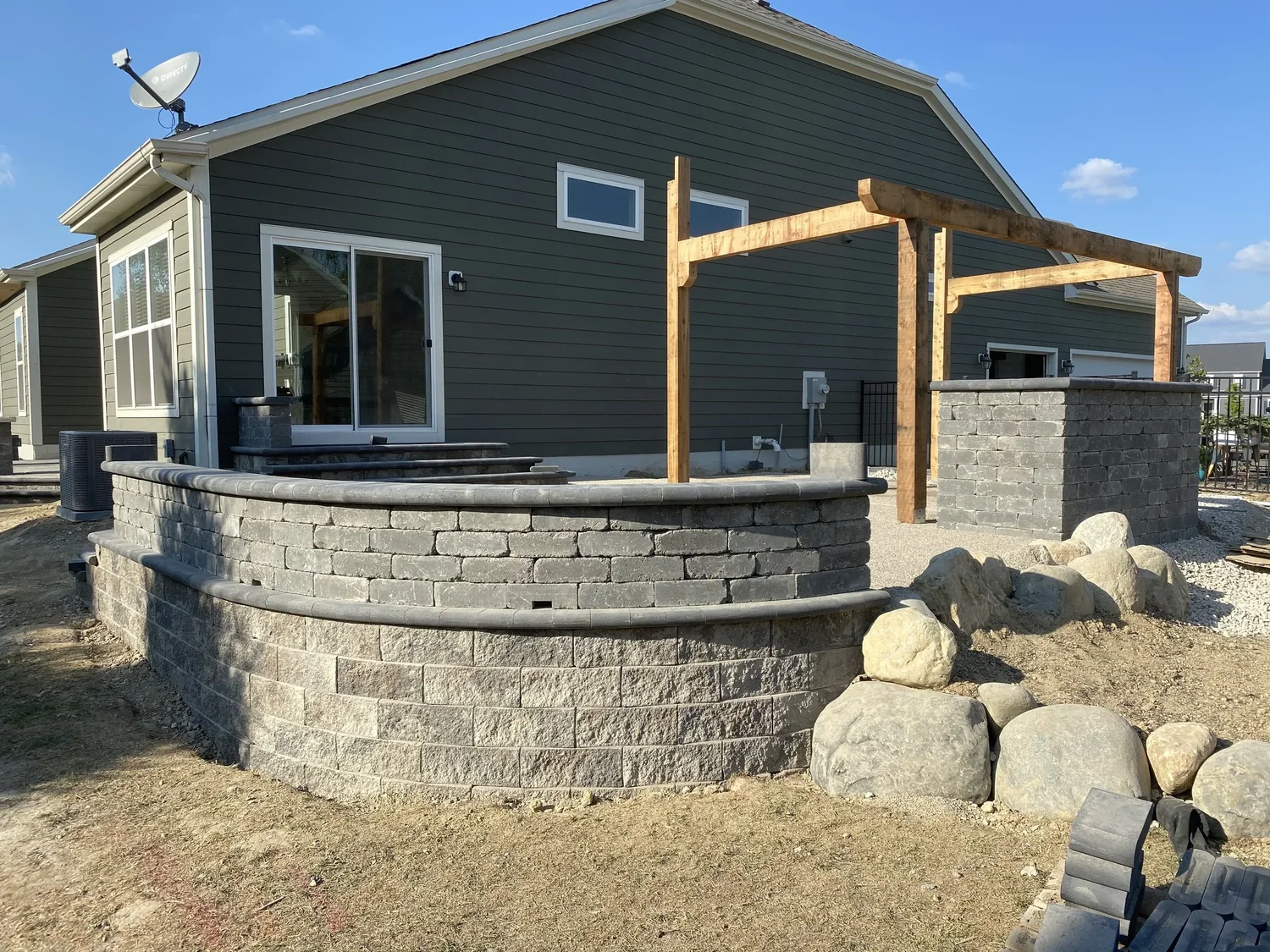 Curved charcoal segmental-block wall with wood pergola posts wrapping the back of a dark-green home in Forsyth County, GA