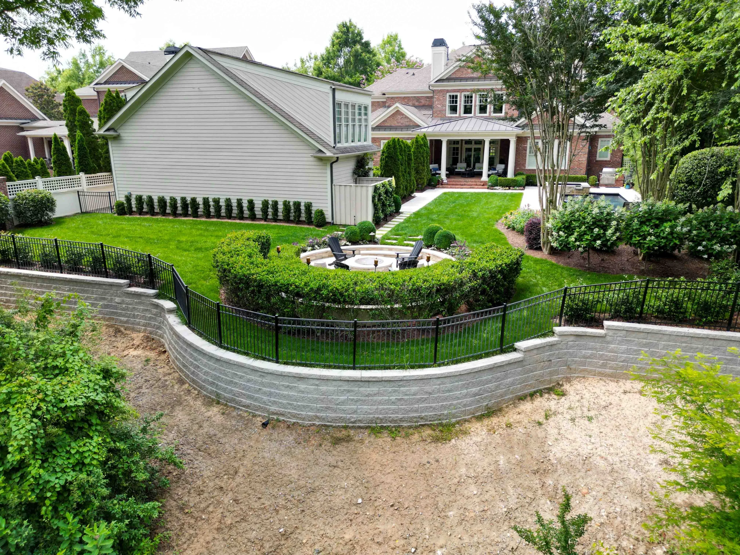 Aerial view of long curved charcoal-block retaining wall along property line behind backyard with iron fence and fire pit in Suwanee, GA