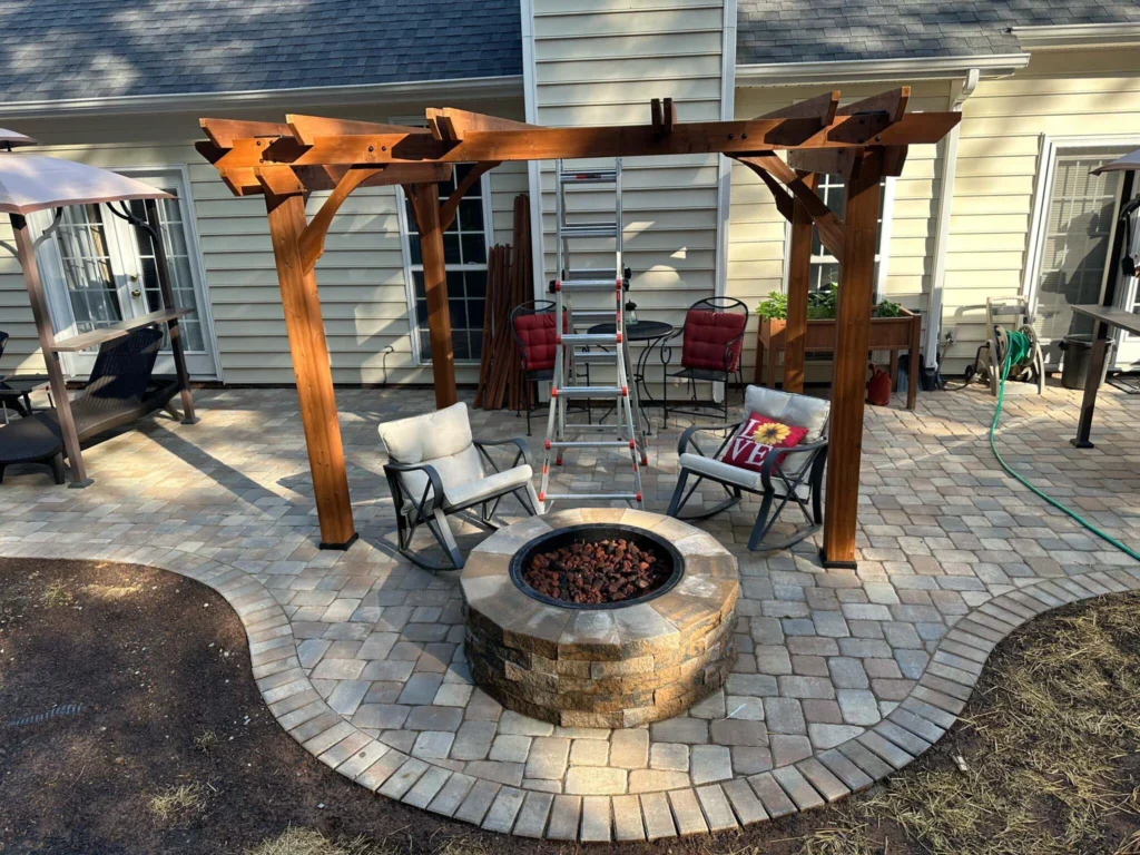 Narrow cedar arbor with octagon-cut beams attached to white siding home, round firepit with red lava rock, 2 padded rockers — backyard firepit and fireplace in Cumming, GA by Primetime Pools.