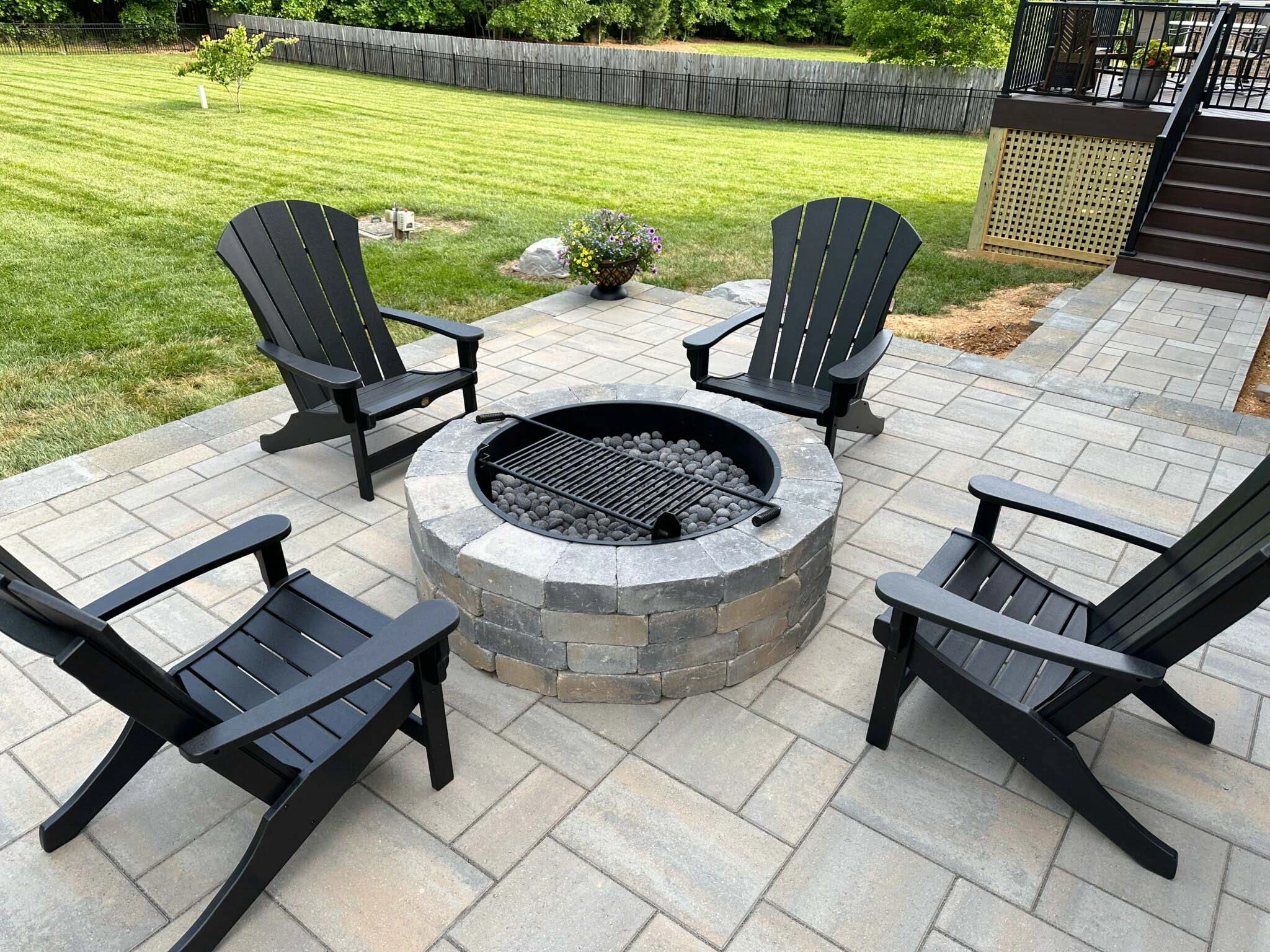 Round stacked-stone gas fire pit with river-rock media and four black Adirondacks on a large-format gray paver patio in Forsyth County, GA.
