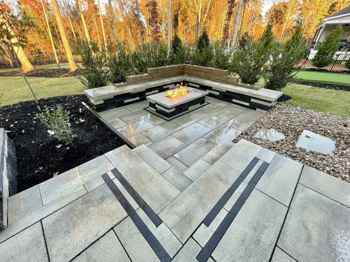 Rectangular linear gas firepit with stacked stone base and bluestone cap surrounded by U-shaped stone bench in wooded autumn Marietta, GA backyard