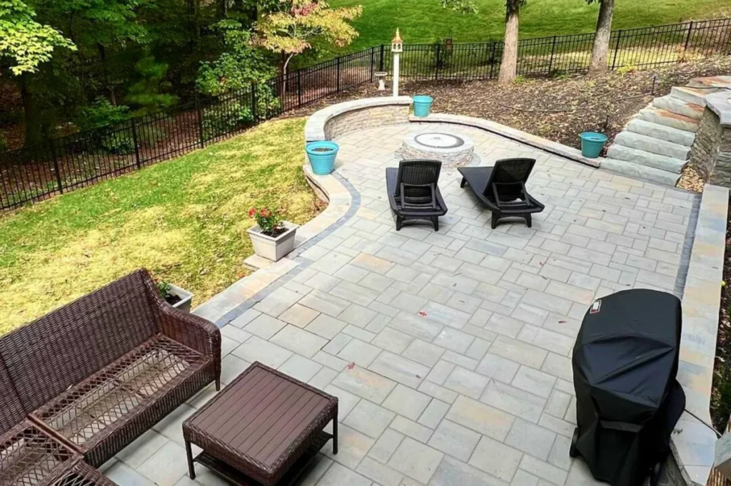 Multi-level gray paver patio with dark wicker sectional, step-up upper tier with modern gray chairs, wooded lot — backyard firepit and fireplace in Milton, GA by Primetime Pools.